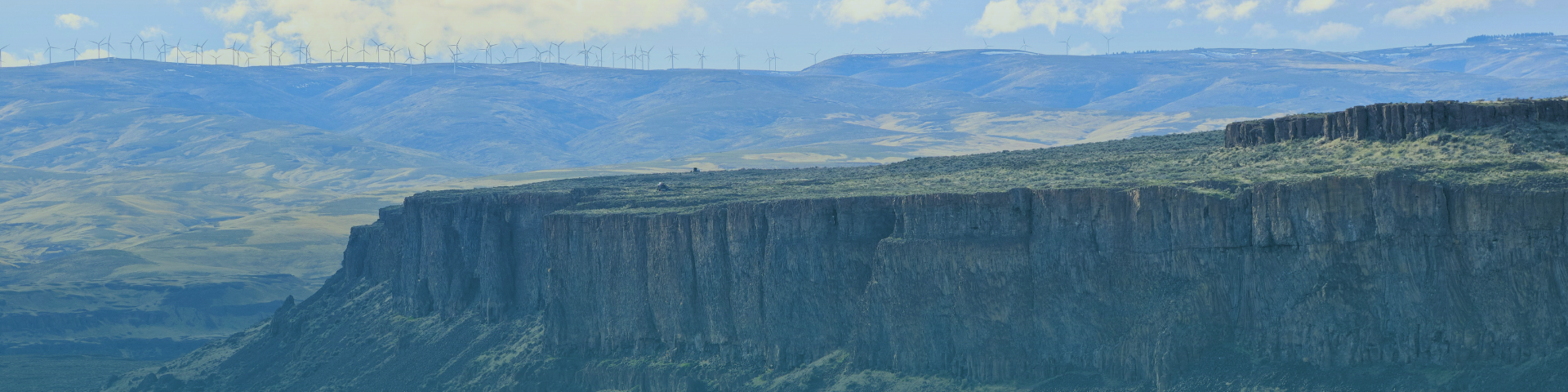 A panoramic view of a vast, rugged landscape featuring a prominent, sheer cliff face in the foreground and rolling hills dotted with wind turbines in the distance under a cloudy sky.