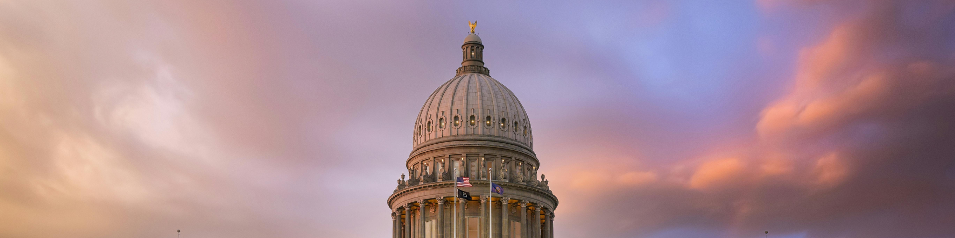 The Idaho State Capitol building with a dome and columns, set against a dramatic sunset sky with pink and purple clouds.